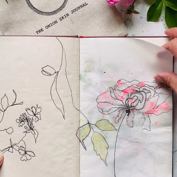 Open sketchbook with floral drawings and a pink flower on a textured surface.