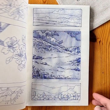 Open book with blue and white illustrations on a wooden surface, with a hand holding the book open.