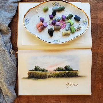 Open book with a painted landscape on one page and a decorative dish with small stones on the other, all on a wooden surface.
